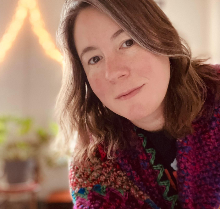 A fair-skinned woman with shoulder-length brown hair and a slight smile. She is wearing a colorful crocheted cardigan. The background is blurred and features a string of lights and a plant.