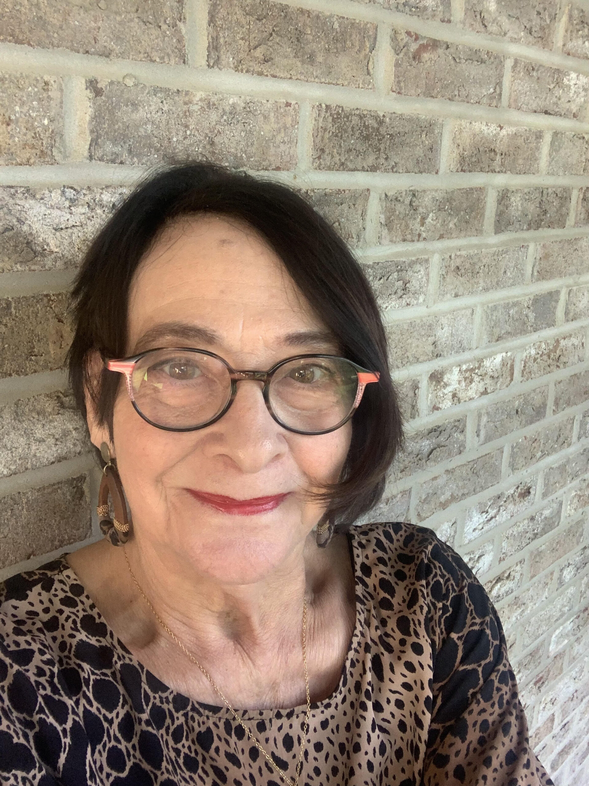 A woman with short, dark hair and glasses smiles at the camera. She is wearing a leopard print top and large, brown earrings. A brick wall is visible behind her.